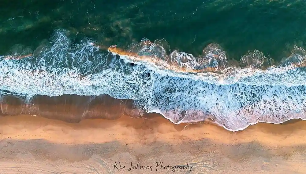 An aerial drone photo shows turquoise ocean waves crashing onto a golden sandy beach, with white foam forming a distinct…