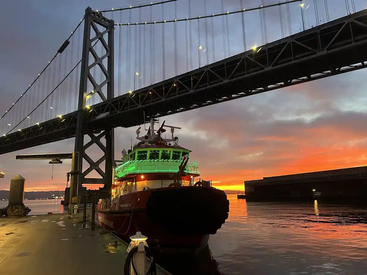A red fireboat named 'FIREBOAT 3' is docked under a large suspension bridge during a vibrant sunset, with orange and pink…