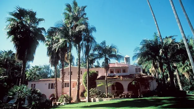 a large, pink stucco house with red-tiled roofs and arched doorways is surrounded by tall palm trees under a clear blue sky…