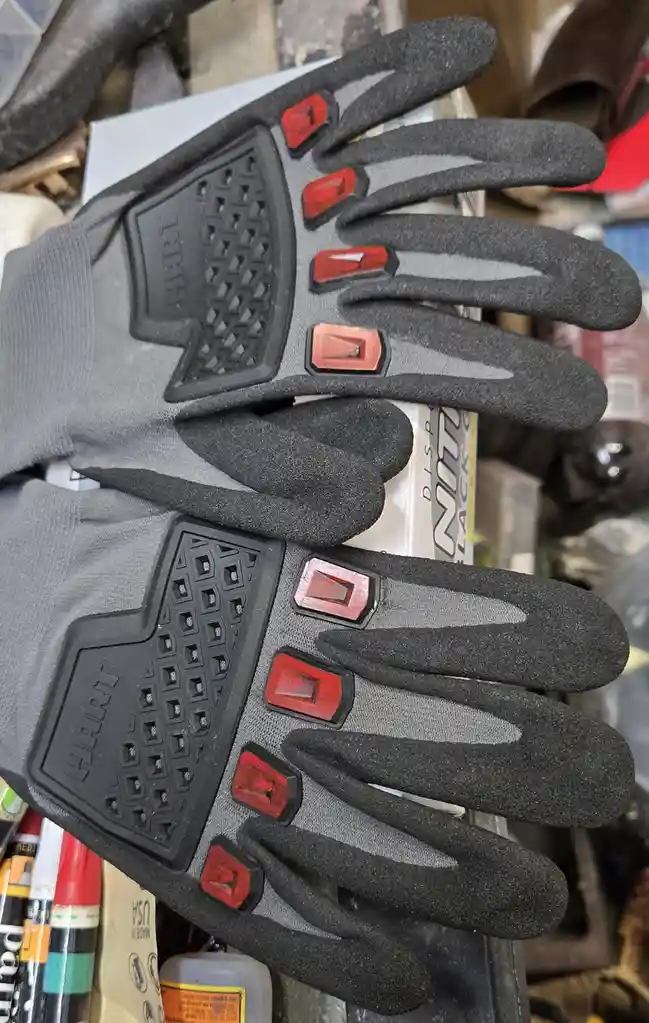 A pair of gray and black work gloves with red rectangular accents on each finger, resting on a cluttered surface with…