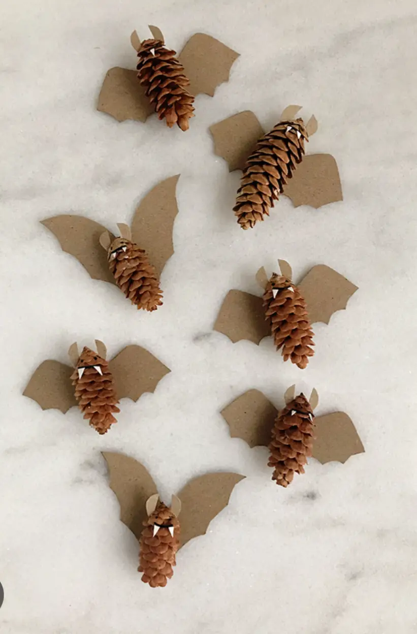 Seven handmade bats made from pinecones with brown paper wings and small white triangular teeth are arranged on a white…