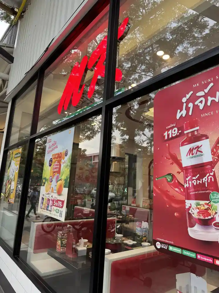 An exterior shot of an MK Restaurants storefront in Thailand, featuring large glass windows displaying red branding and…