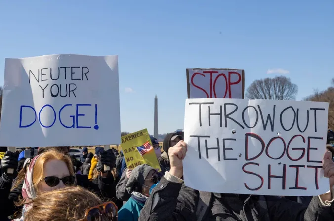 A crowd of protesters holds signs at a rally with the Washington Monument visible in the background. One sign reads 'NEUTER…