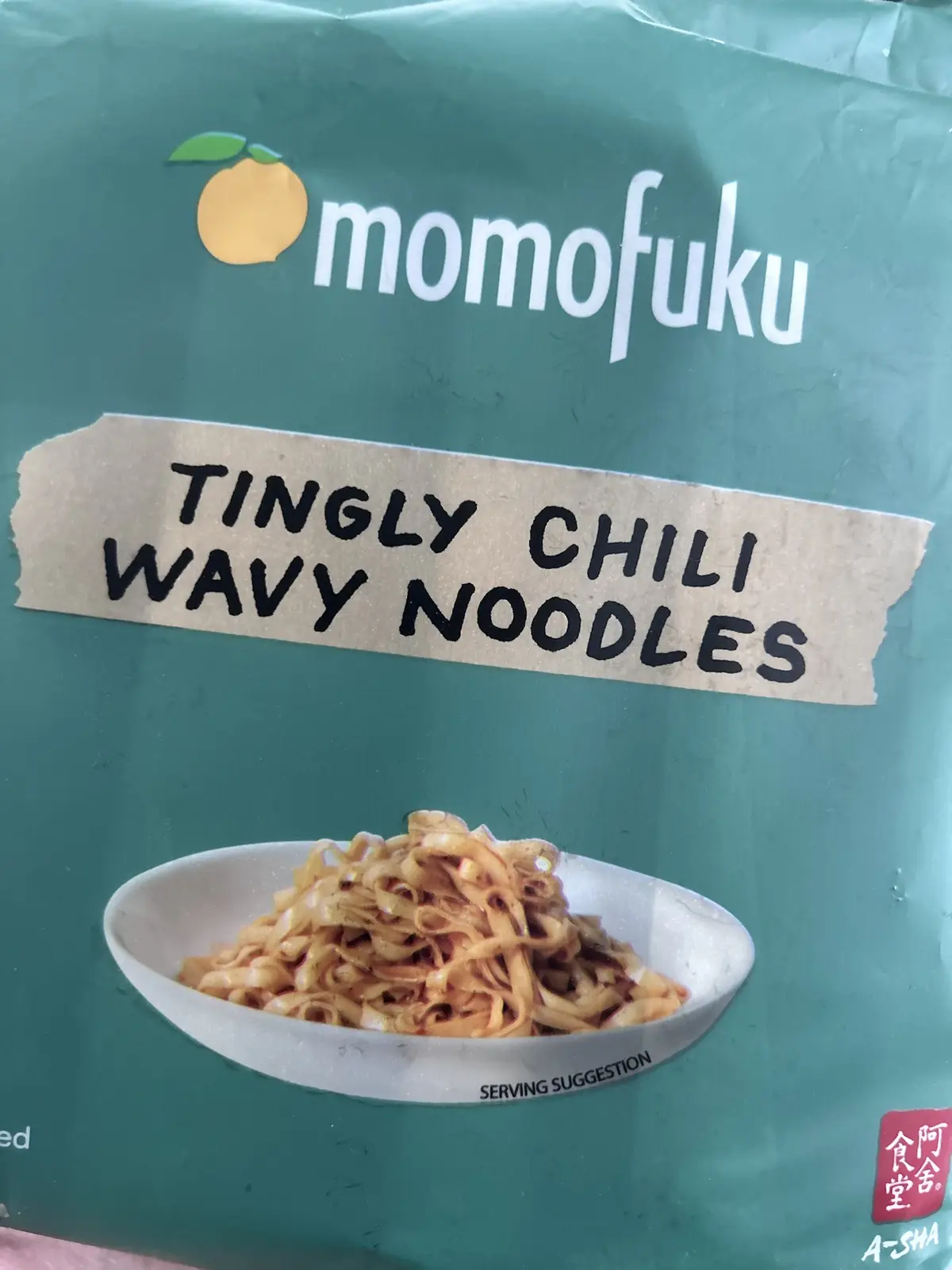 A close-up of a green Momofuku noodle package showing a bowl of wavy noodles. A piece of brown paper tape is overlaid on the…