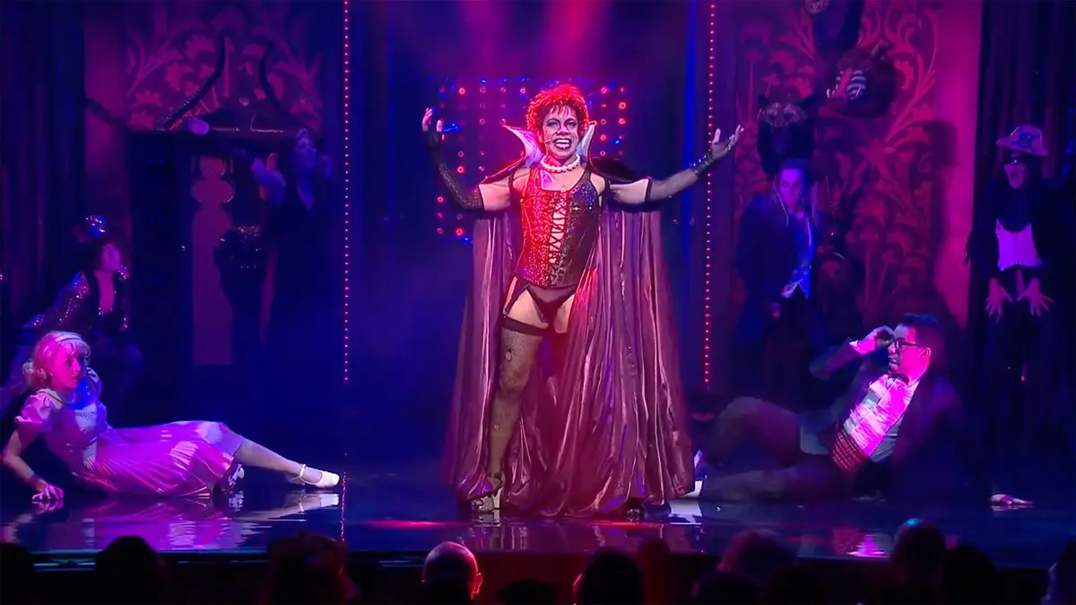 A performer in a red devil costume with horns and a cape stands center stage with arms outstretched, smiling broadly. To…