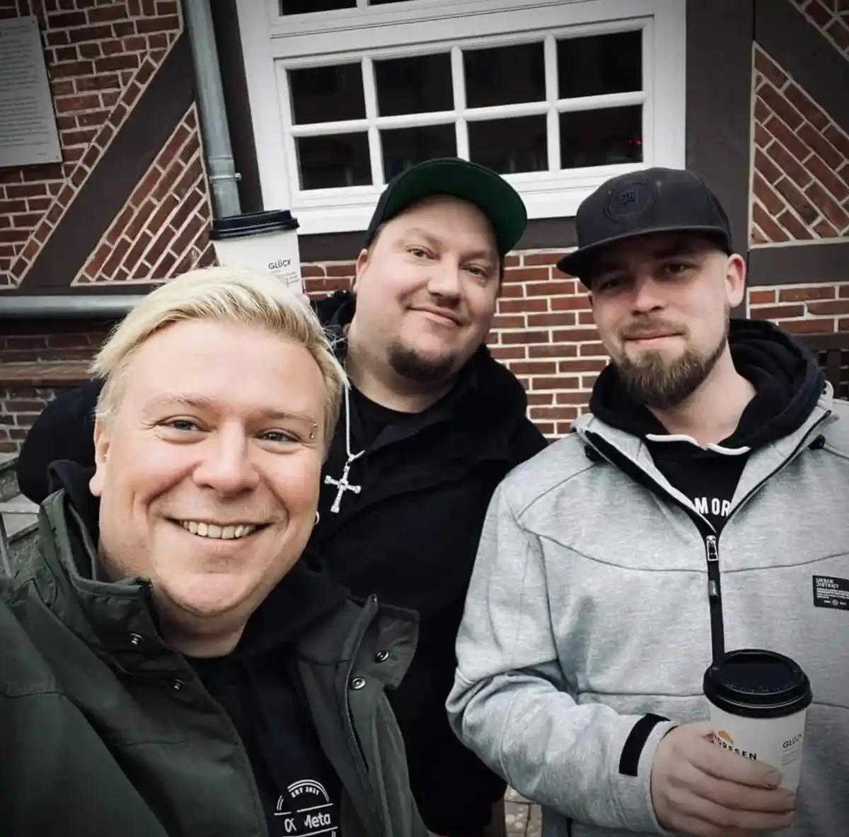 Three men pose for a selfie outdoors in front of a brick building with a white window. The man on the left has blonde hair…