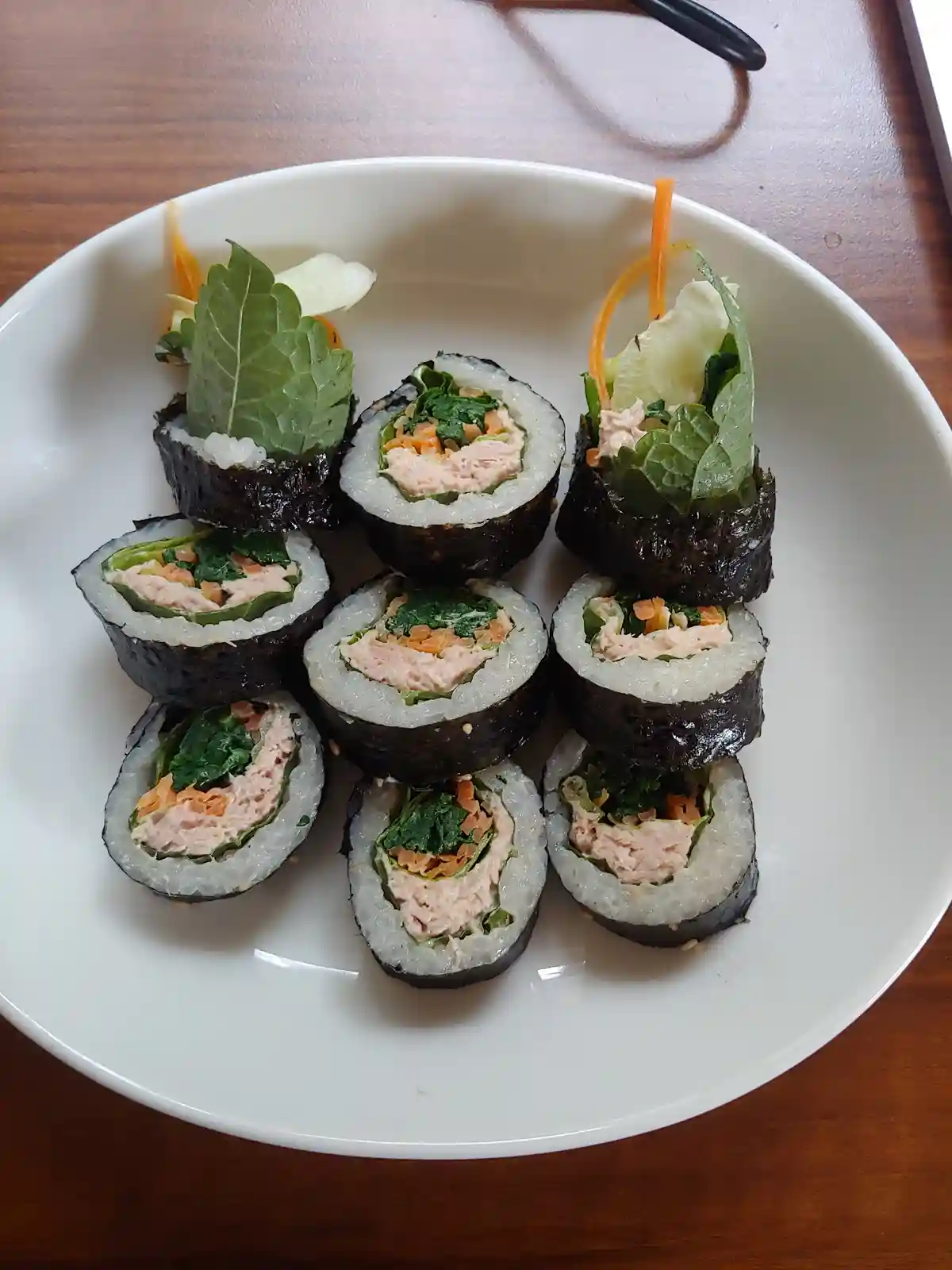 A white plate holds eight neatly arranged sushi rolls, each cut in half to show pink filling, green leaves, and orange…