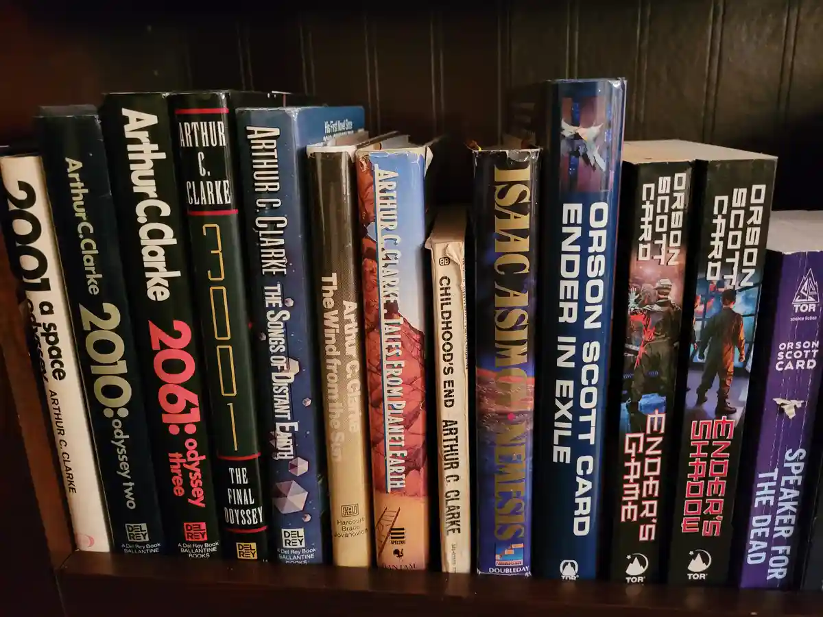 A close-up shot of a wooden bookshelf filled with science fiction novels, primarily by Arthur C. Clarke, Isaac Asimov, and…