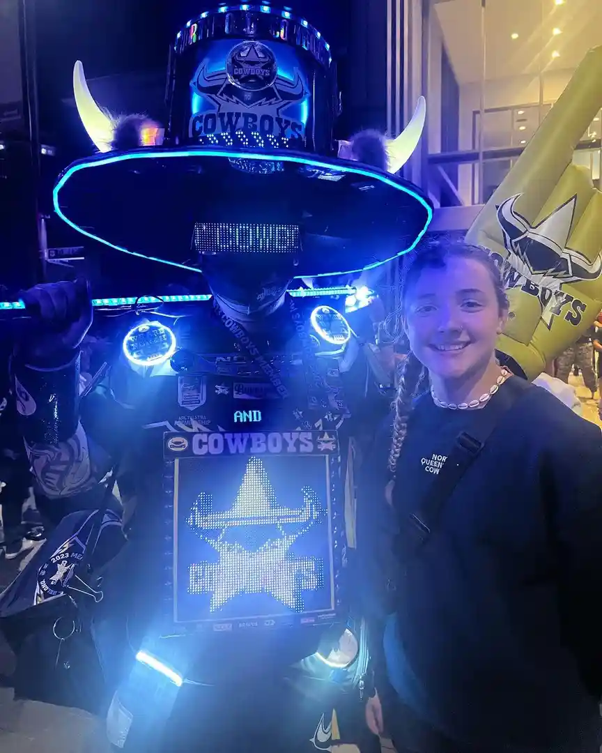 A person in an elaborate, glowing blue costume with a large cowboy hat, LED lights, and a 'COWBOYS' logo stands next to a…