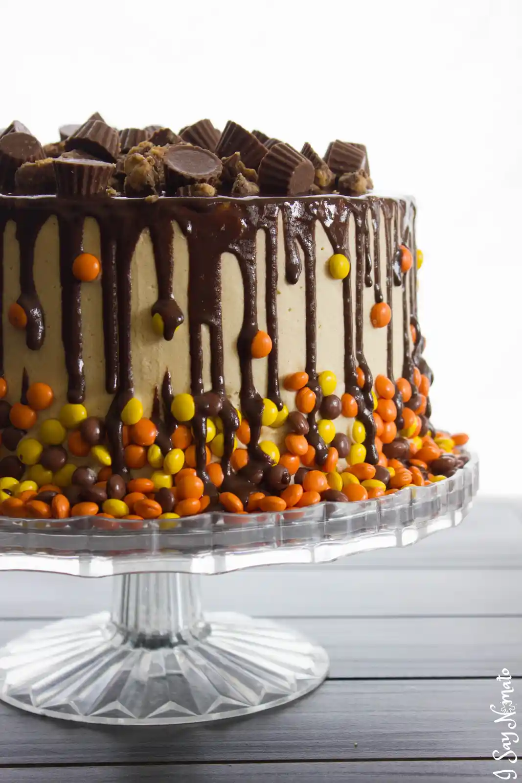 A close-up of a cake with light brown frosting, chocolate drizzle, and topped with crushed peanut butter cups and colorful…