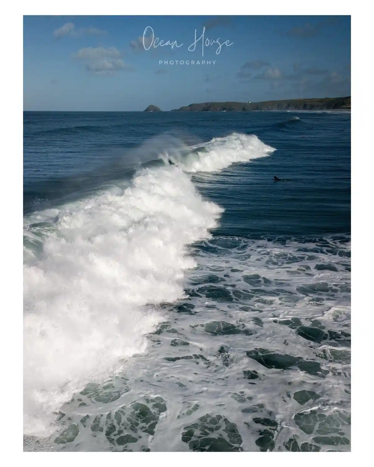 A photograph of powerful ocean waves crashing with white foam, showing a surfer in the distance and a rocky coastline under…