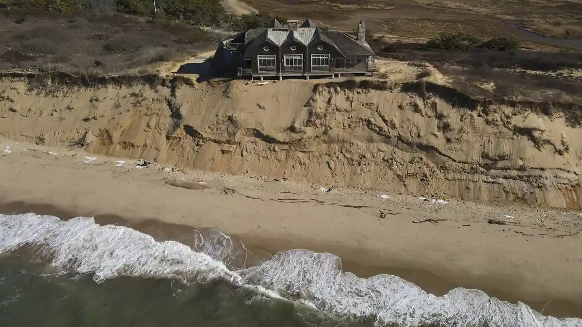 An aerial photo shows a large house perched precariously on the edge of a severely eroded cliff overlooking a sandy beach…