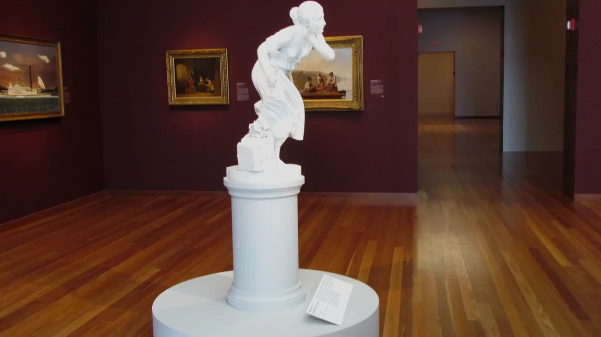 A white marble statue of a woman in a flowing gown, posed as if weeping, stands on a pedestal in a museum gallery with dark…