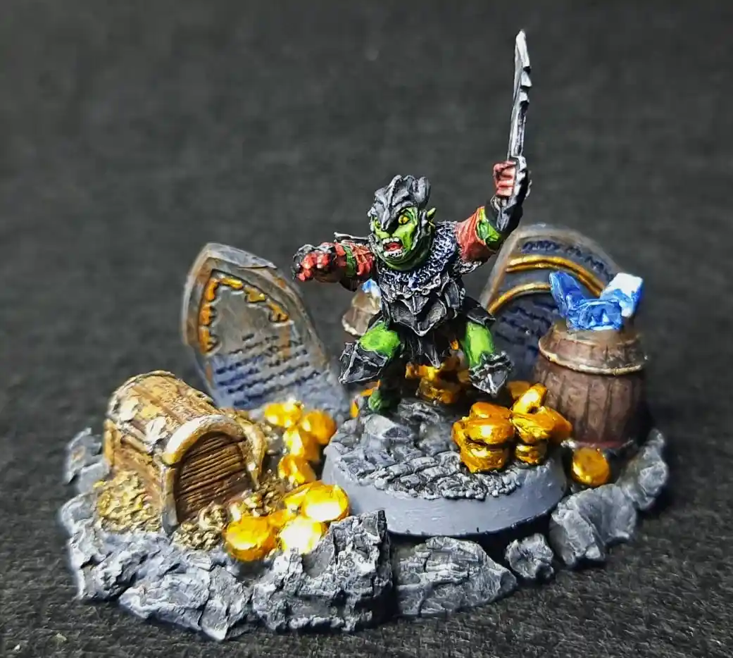 A painted miniature of a green-skinned orc-like creature in armor, holding a sword aloft, stands triumphantly on a rocky…