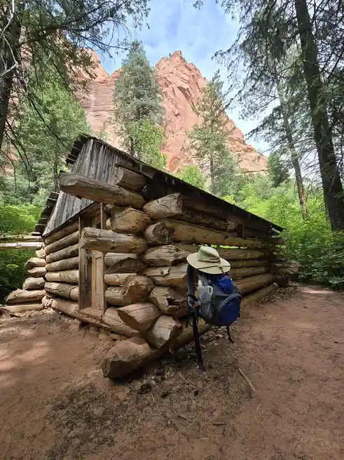 A rustic log cabin sits in a forest with red rock formations in the background. A hiking backpack with a wide-brimmed hat…