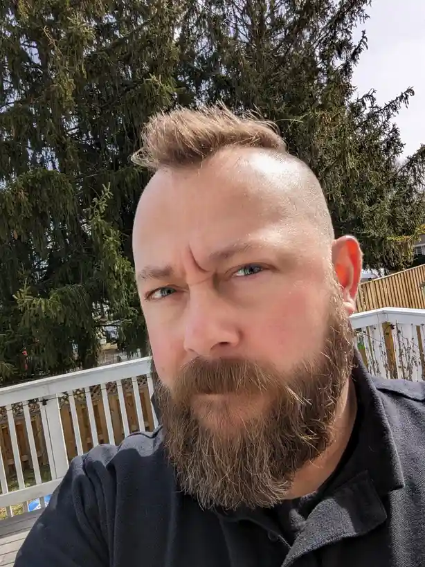 A man with a shaved head and full beard makes a scowling, skeptical face at the camera while standing on a wooden deck with…