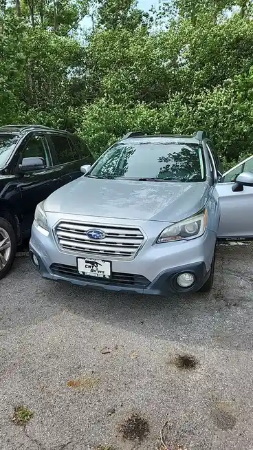 A silver Subaru Outback is parked outdoors with its driver's side door open, next to a black SUV. The car's front grille and…