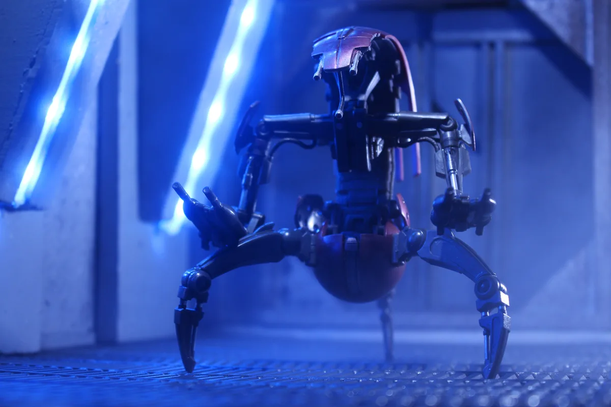 A small, spider-like robot toy with black and red parts stands on a textured metal floor under blue lighting, with glowing…