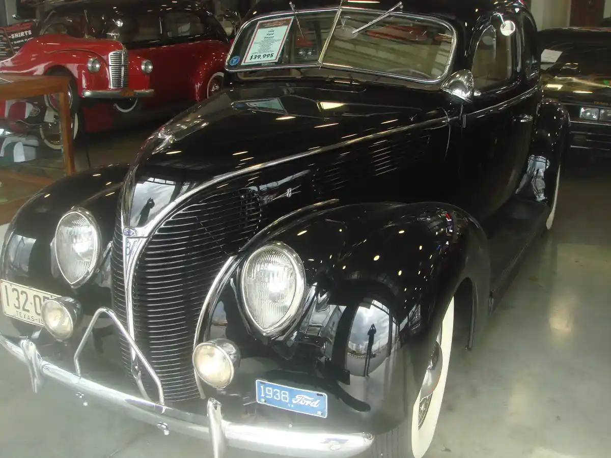 A glossy black 1938 Ford Deluxe is displayed in a car showroom, with a price tag of $39,995 visible on the windshield. The…