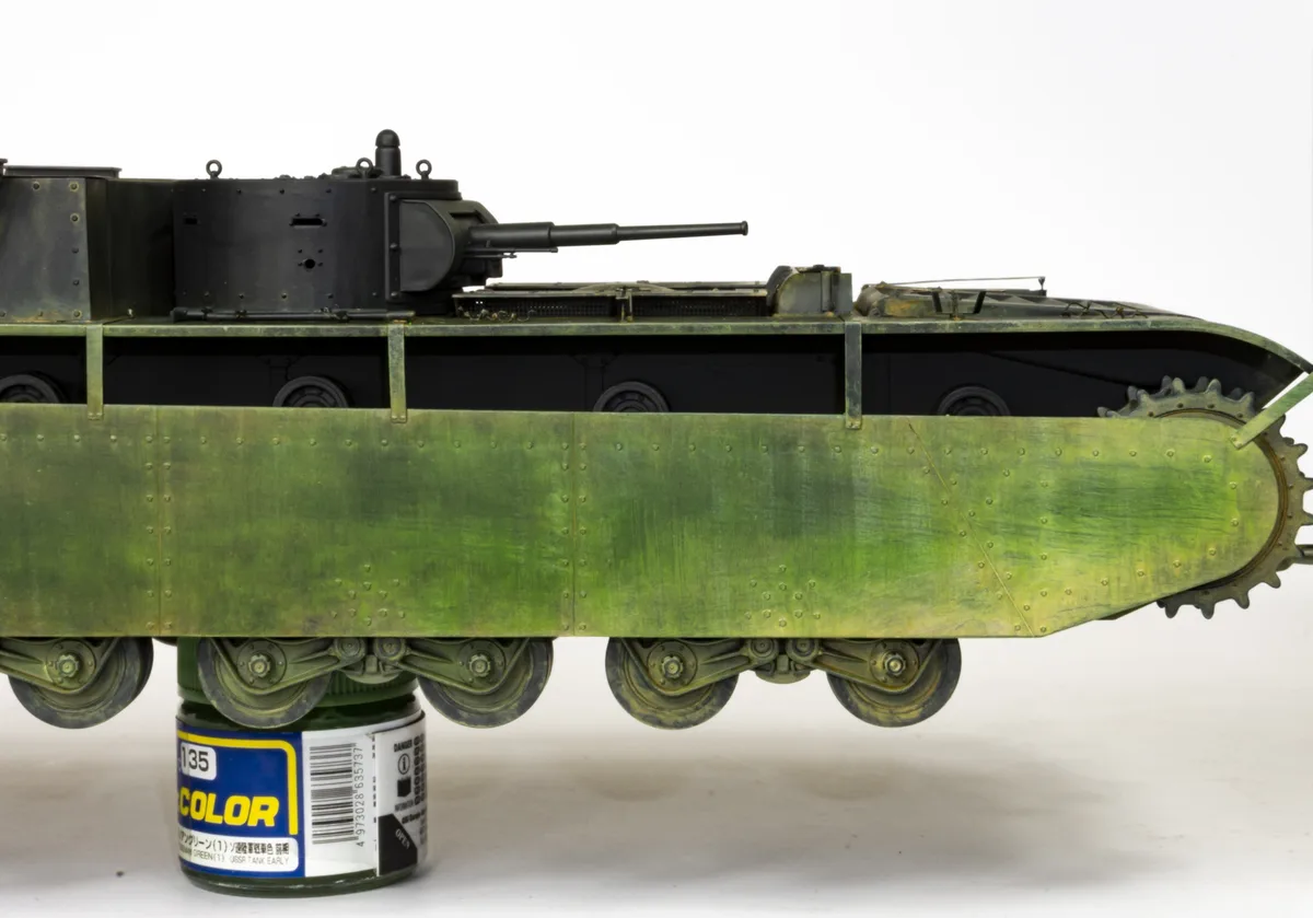 A scale model of a green military tank is shown resting directly on top of a green paint can, with the tank's undercarriage…