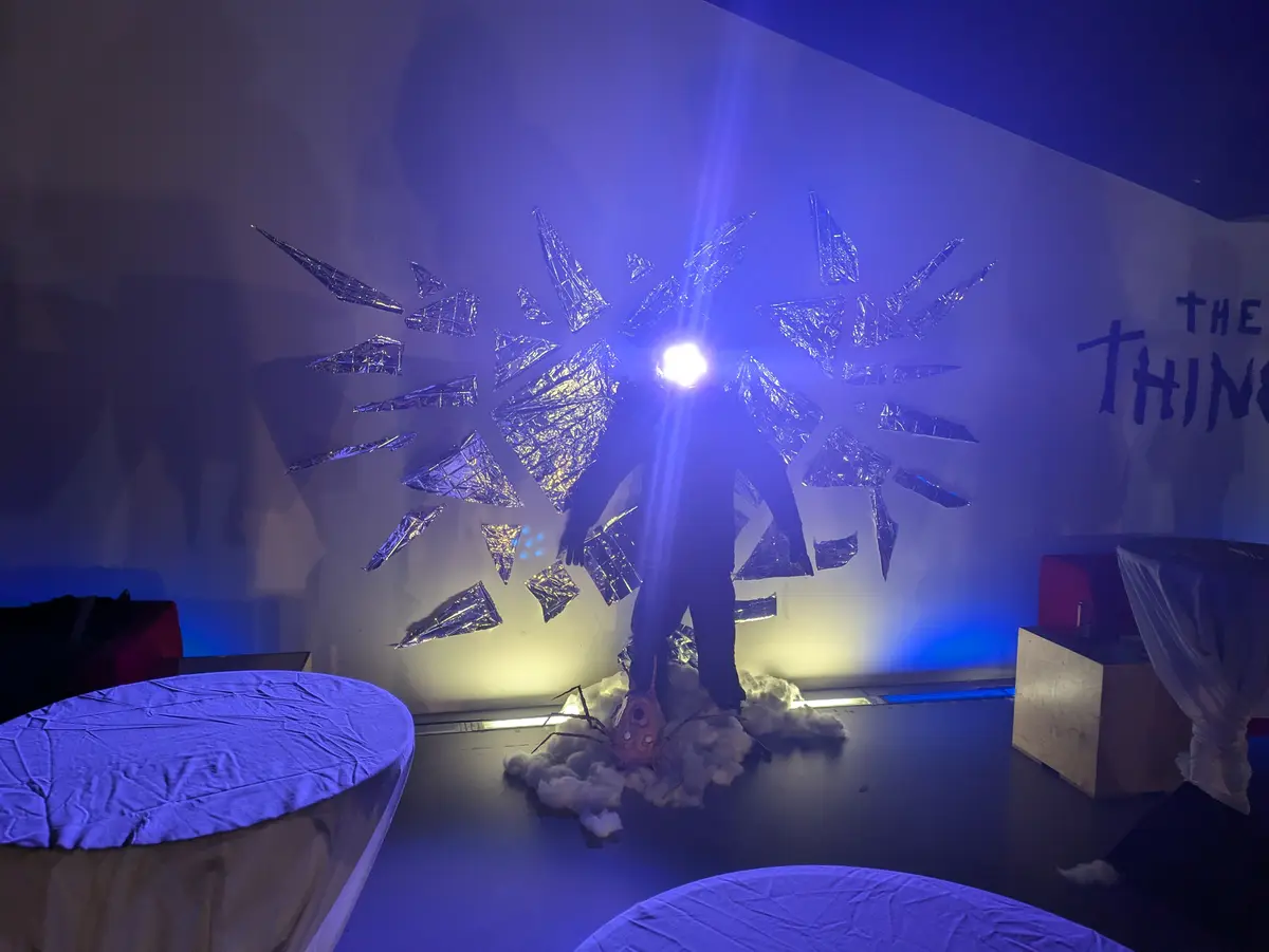 A person stands in silhouette on a stage bathed in blue light, surrounded by a burst of shiny, foil-like shards radiating…