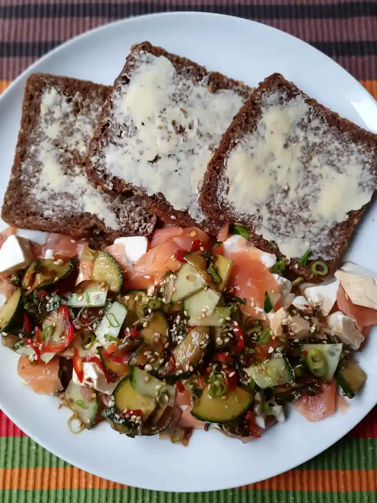 A white plate holds three slices of dark brown bread spread with butter, arranged above a colorful salad of smoked salmon…