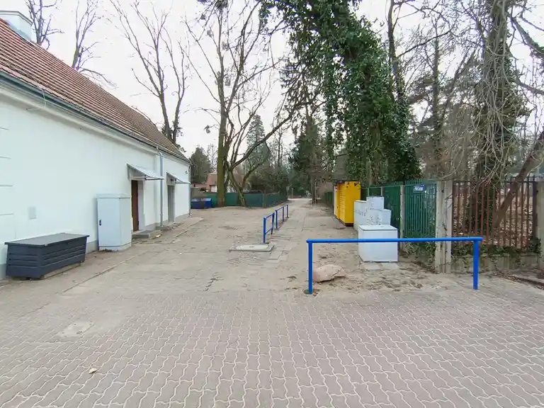 An outdoor courtyard with a paved path, a white building on the left, and various utility boxes and blue railings. A large…