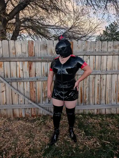 A person stands outdoors in front of a wooden fence wearing a full-body black latex outfit with red accents, including a…