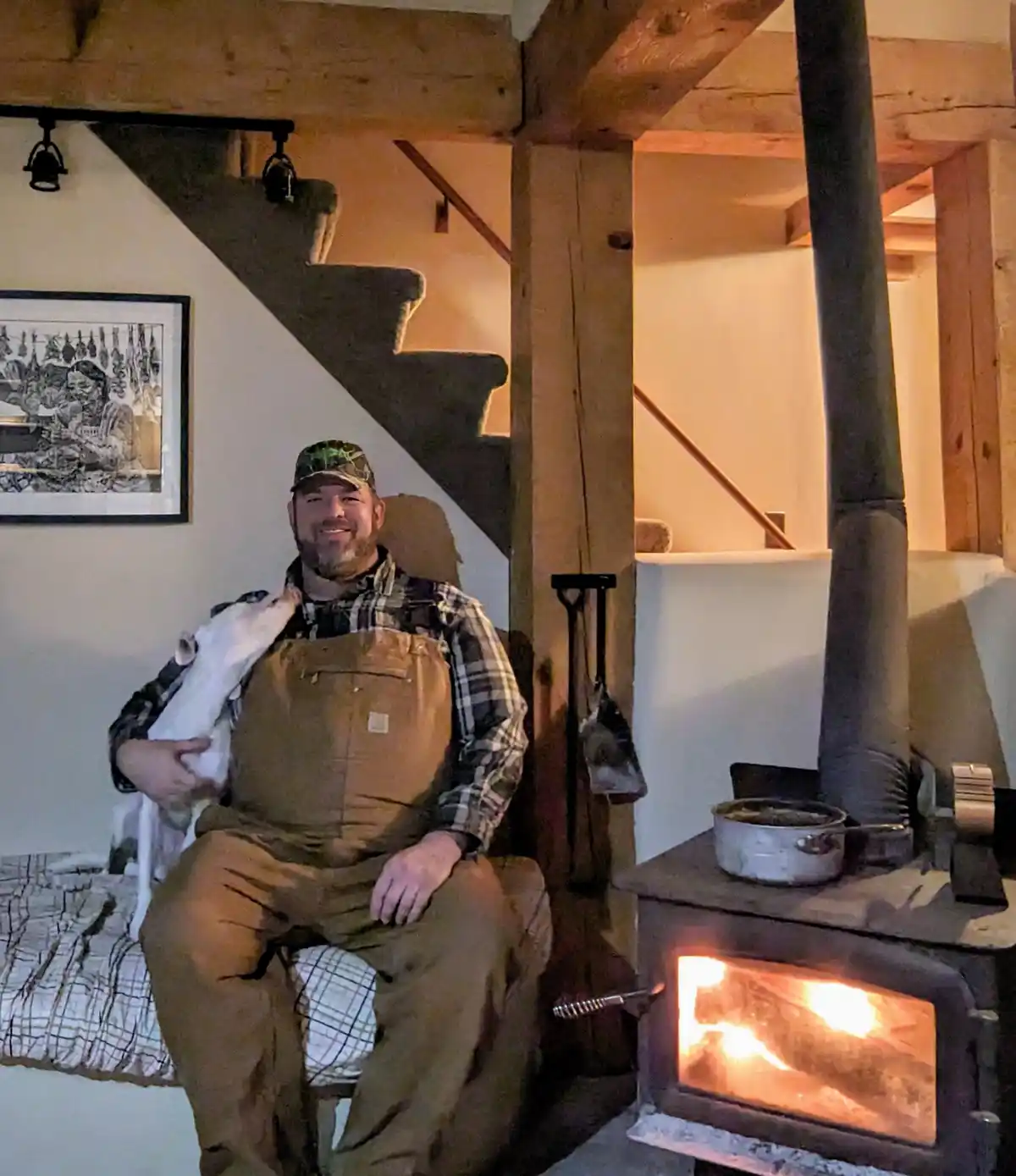 a man in overalls and a camouflage cap sits smiling in a cozy room with exposed wooden beams, holding a white dog. Beside…