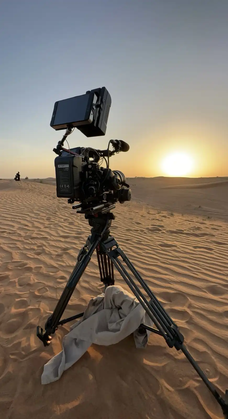 A professional video camera mounted on a tripod is set up in a desert at sunset, with the sun low on the horizon. The camera…