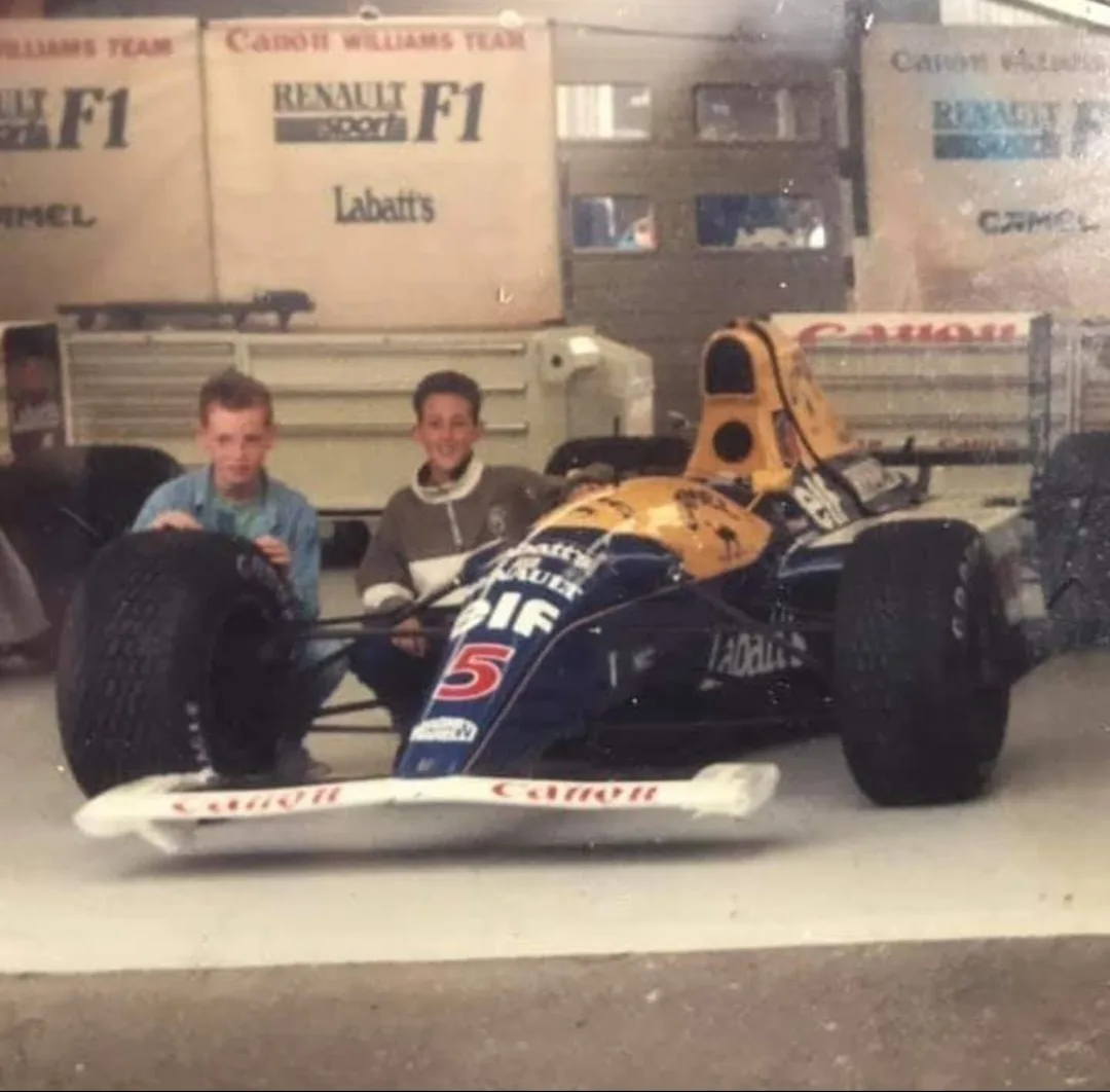 Two young boys are kneeling beside a vintage blue and yellow Formula 1 race car with 'Canon' and 'Renault' branding, in…
