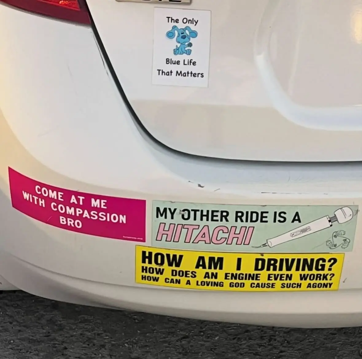 A close-up of a white car's rear bumper and trunk adorned with multiple humorous bumper stickers. One sticker features a…