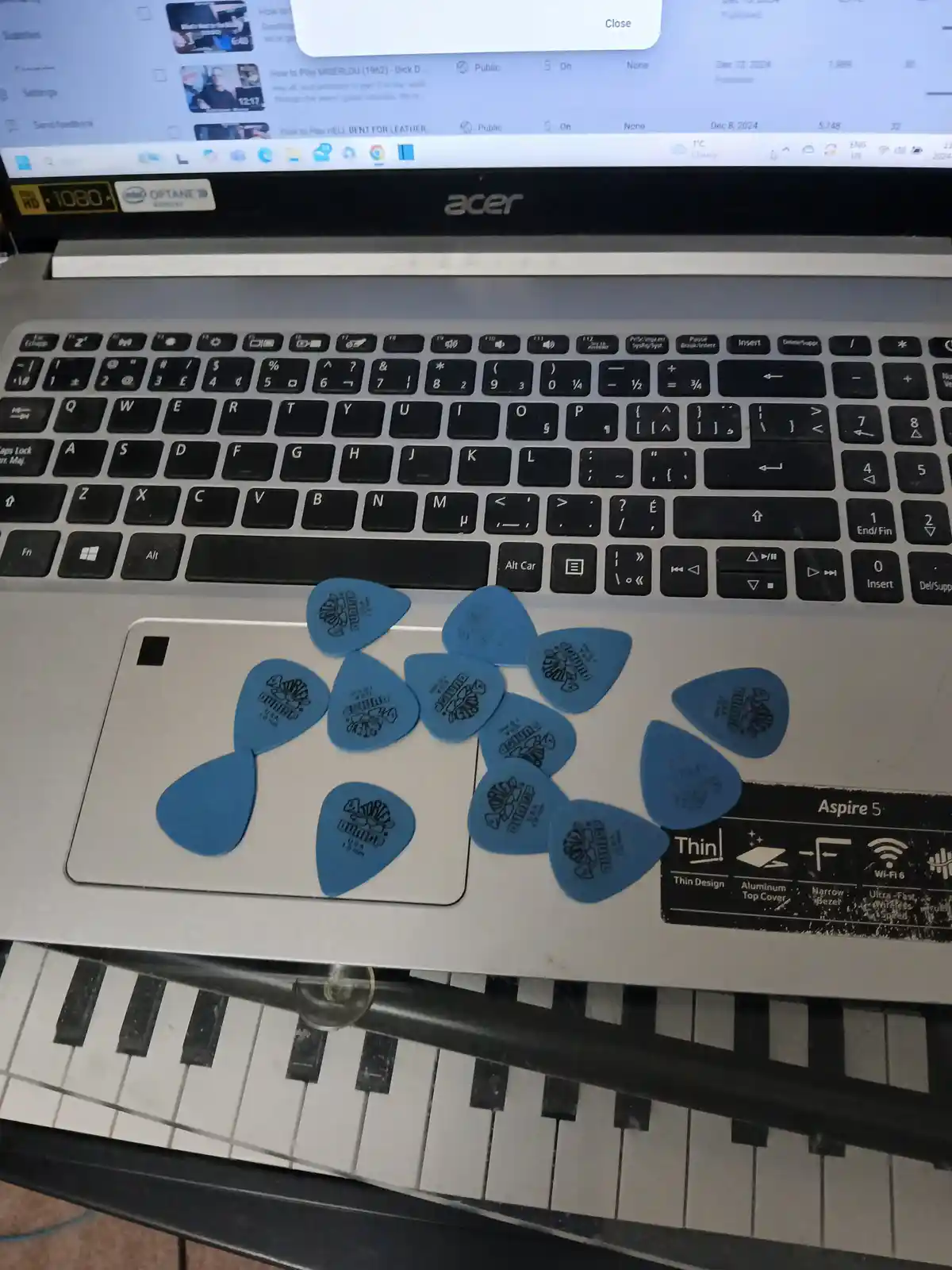 A close-up photo shows numerous blue Dunlop guitar picks scattered across the silver touchpad and keyboard of an Acer Aspire…