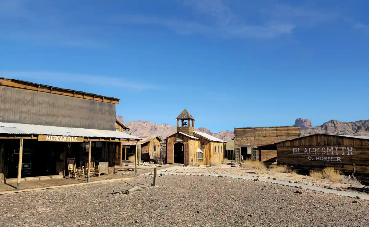 A wide shot of a deserted, sun-baked Western ghost town with wooden buildings labeled 'MERCANTILE', 'CHURCH', 'CASTLE CITY'…