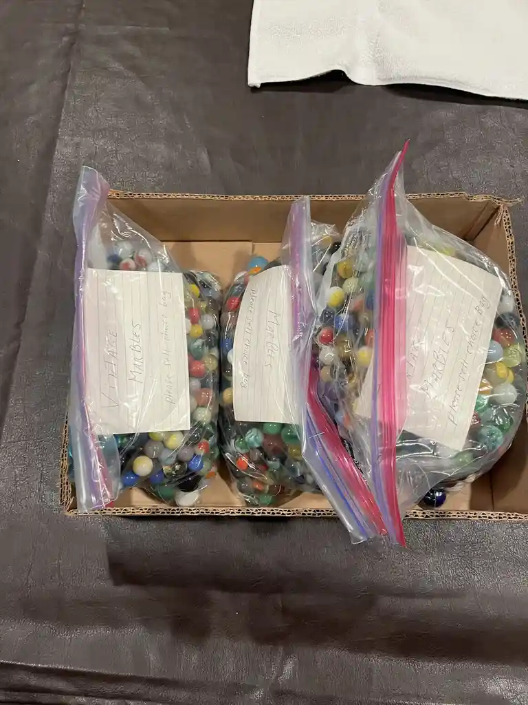 Three zip-top bags filled with colorful marbles sit inside a cardboard box on a dark surface. Each bag has a handwritten…