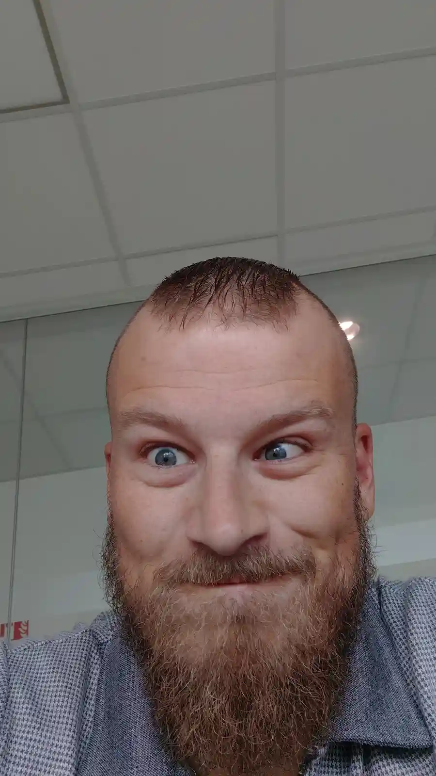 A close-up selfie of a man with a buzz cut and a full beard, wearing a patterned collared shirt. He has wide…