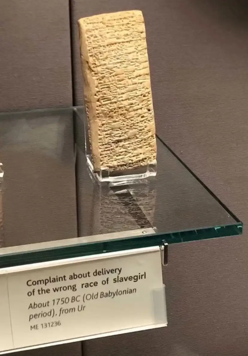 An ancient cuneiform tablet, displayed in a museum case, has an informational plaque below it that reads 'Complaint about…