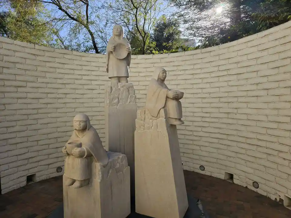 Three stone statues of figures in robes, each holding a bowl or plate, stand on pedestals in a courtyard with a white brick…