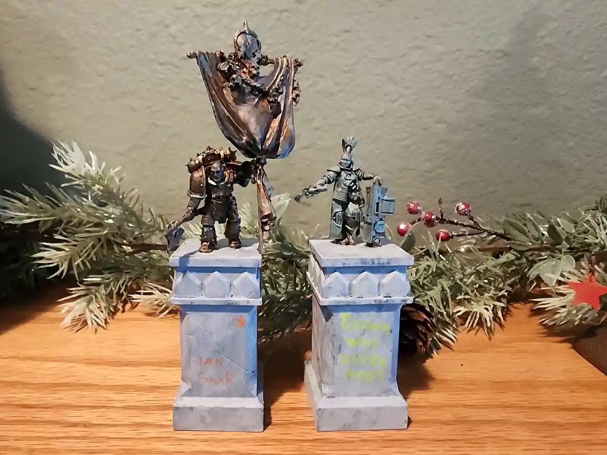 Two painted miniature figurines stand atop small tombstone-like bases on a wooden surface, surrounded by Christmas garland…