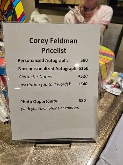 A printed sign titled 'Corey Feldman Pricelist' lists fees for autographs and photo opportunities, including personalized…