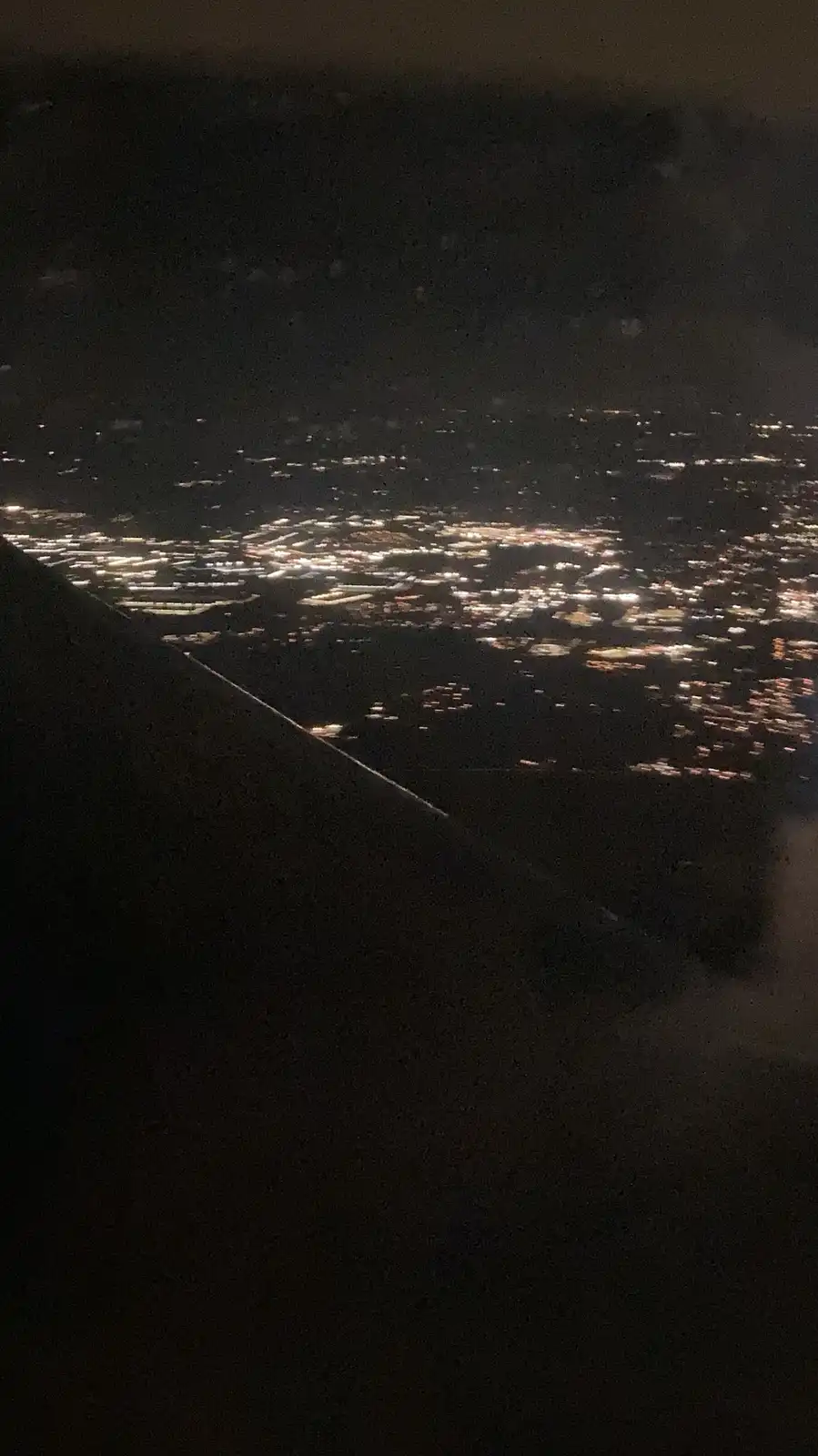 A nighttime photo taken from an airplane window showing the dark silhouette of the wing in the foreground and a sprawling…