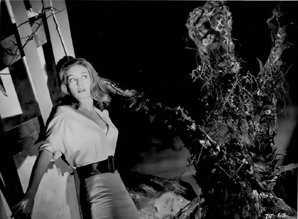 A black-and-white still from a horror film shows a woman in a light blouse and dark pants looking up in terror at a…