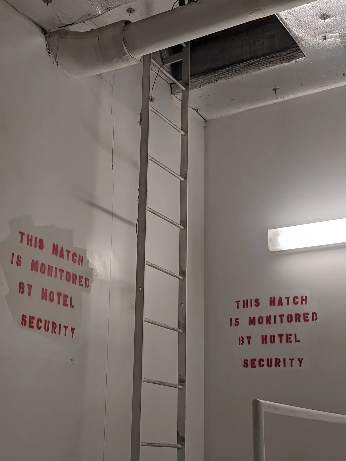 A metal ladder leans against a white wall next to a ceiling hatch, with red stenciled text on both walls reading 'THIS HATCH…