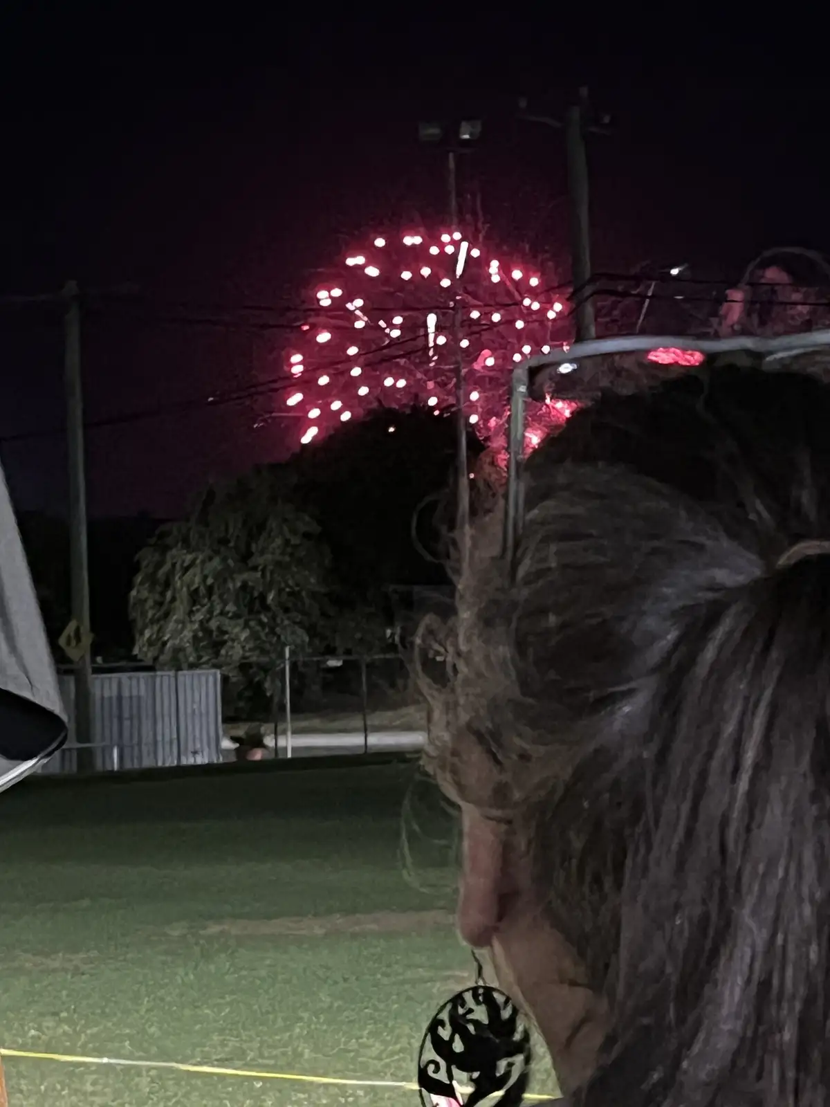 A person viewed from behind is watching red fireworks explode in the night sky, with trees and utility poles visible in the…