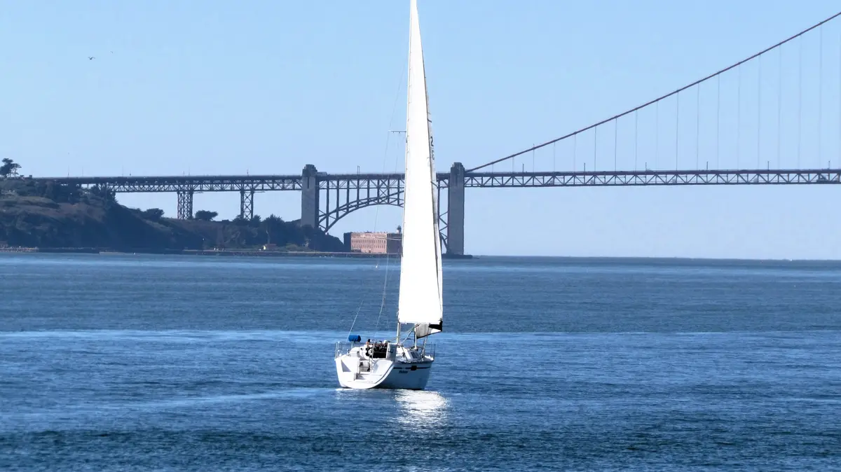 A white sailboat with a large white sail glides across calm blue water under a clear sky, with a large suspension bridge…