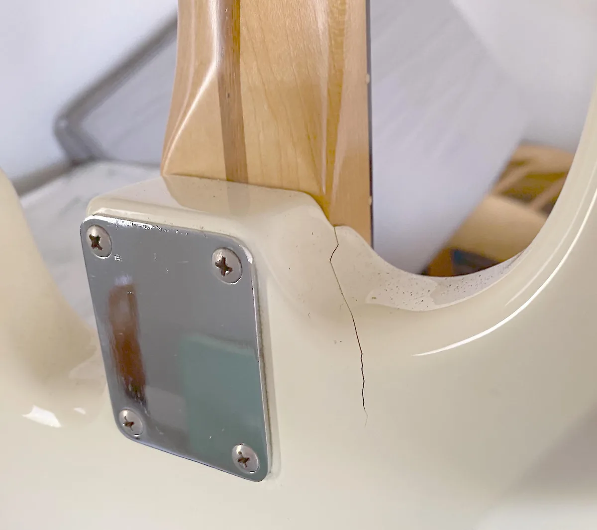 A close-up photo shows a small, dark crack running from the edge of a cream-colored guitar body near the neck joint. A…