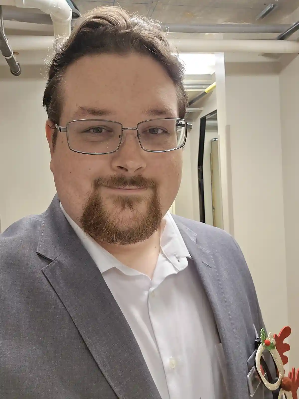 A man with glasses and a beard, wearing a grey suit jacket over a white shirt, stands in an office hallway with exposed…