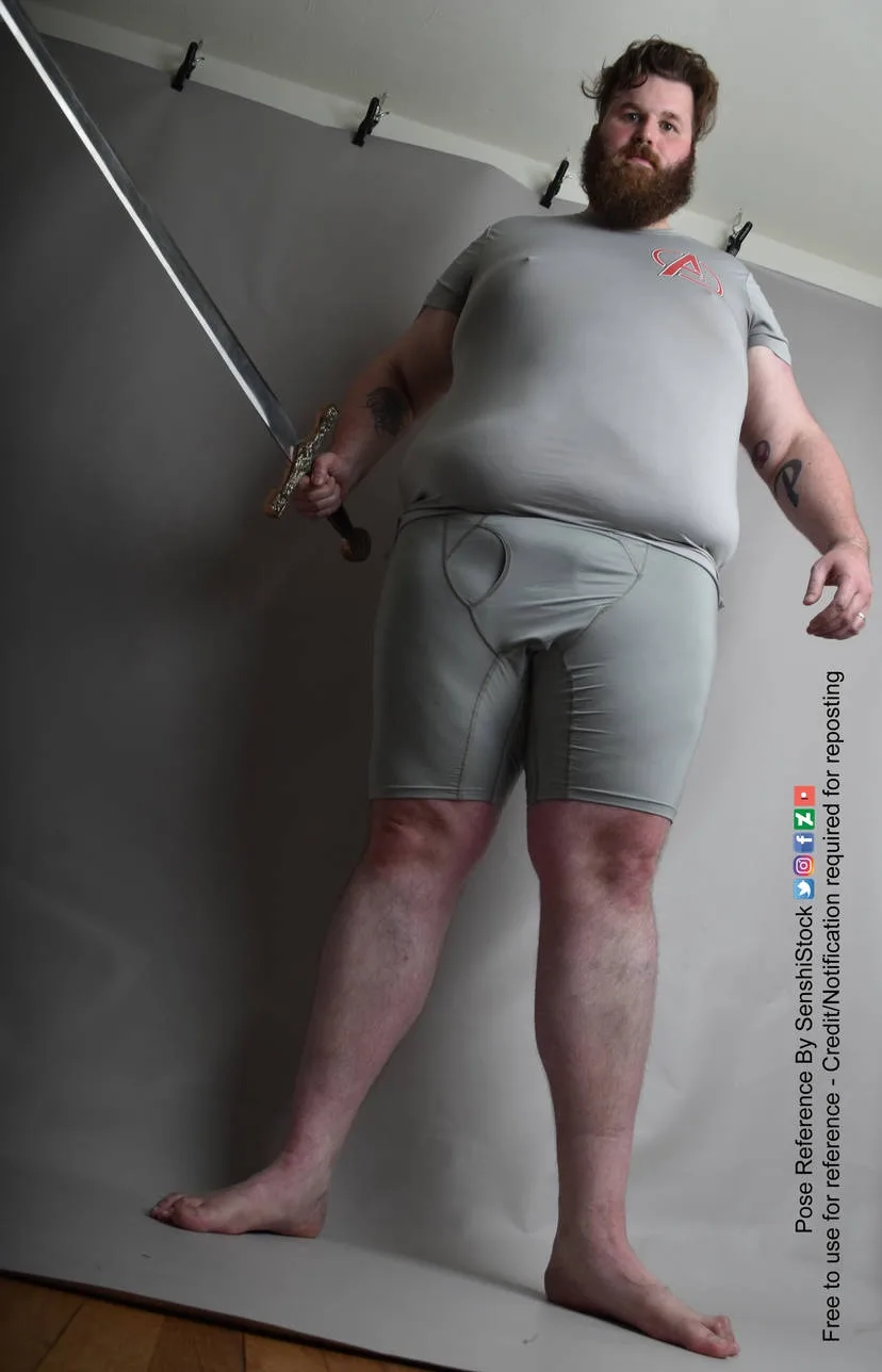 a man with a beard and tattoos stands barefoot on a gray backdrop, holding a sword in his right hand, wearing a gray t-shirt…
