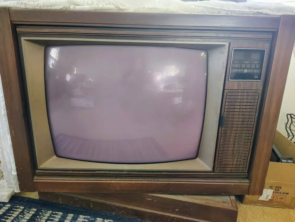An old wooden-framed General Electric CRT television with a blank, slightly purplish screen, sitting on a stand. The screen…