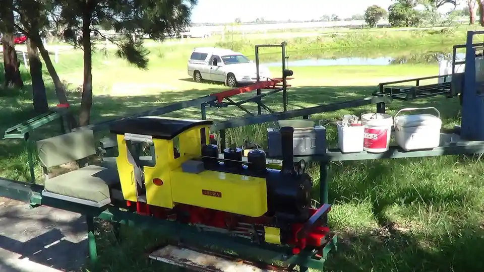 A yellow and black miniature steam train named 'Brenda' sits on a green metal track outdoors, with a white pickup truck…